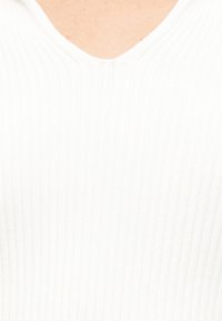 Personne portant un pull à col en V côtelé blanc, montrant le haut de la poitrine et l'encolure sur un fond uni.