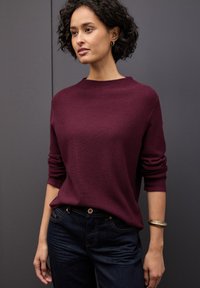 Burgunderfarbener Strickpullover mit Rundhalsausschnitt und aufgeschlagenen Ärmeln, kombiniert mit dunkelblauen Jeans und einem strukturierten goldenen Armband.