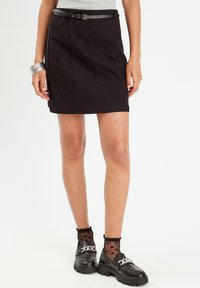 Jupe noire avec une finition texturée, dotée d'une fente latérale et d'une ceinture en cuir fine. Associée à des mocassins noirs et des chaussettes à motifs.