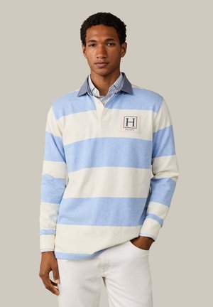 Hombre vestido con camisa de rugby de manga larga con rayas horizontales en azul claro y blanco con cuello, pantalones blancos, de pie con una mano en el bolsillo.