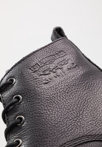 Bota de cuero negro con superficie texturizada, equipada con ojales de metal y cordones oscuros. Detalle del logo embellecido en el costado.