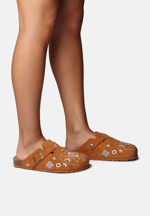 Zoccoli in camoscio marrone con motivi ricamati in blu, bianco e giallo, caratterizzati da una fibbia decorativa e da una suola in gomma con texture.