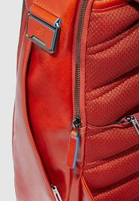 Rode leren en stoffen rugzak met een gestructureerd oppervlak, voorzien van een ritssluiting, metalen hardware en accentdetails in het blauw.