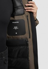 Kabát s čiernou polstrovanou podšívkou a khaki vonkajšou stranou so zipsom, zdobený nášivkou s logom a hladkým, textúrovaným materiálom.