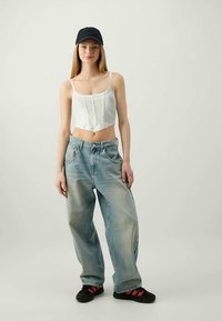 Weißes, kurzes Oberteil, hellblaue, weite Jeans und schwarze Mütze. Das Model trägt schwarze Schuhe mit roten Akzenten. Einfaches Design, lässige Passform und weicher Stoff.