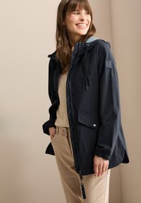 Chaqueta impermeable navy con puños elásticos, capucha con cordón y bolsillos frontales, combinada con una blusa beige clara y pantalones beige.