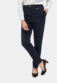 Marineblauwe broek van stof met een gladde textuur, voorzien van een gouden knoop, riemlussen en een getailleerde pasvorm; gecombineerd met zwarte hakken.