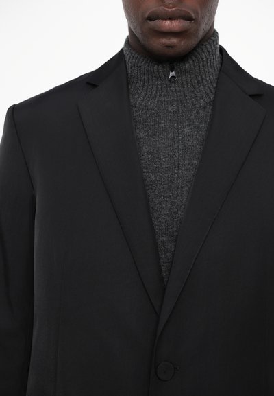 Blazer negru tailored cu rever încrețit, purtat peste un pulover tricotat de culoare gri închis, cu guler înalt. Material neted, închidere cu un singur nasture și design minimalist.