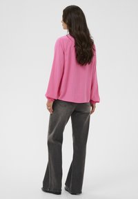 Femme aux longs cheveux noirs portant un blouse rose à manches longues et un jean noir ample, se tenant contre un fond clair.