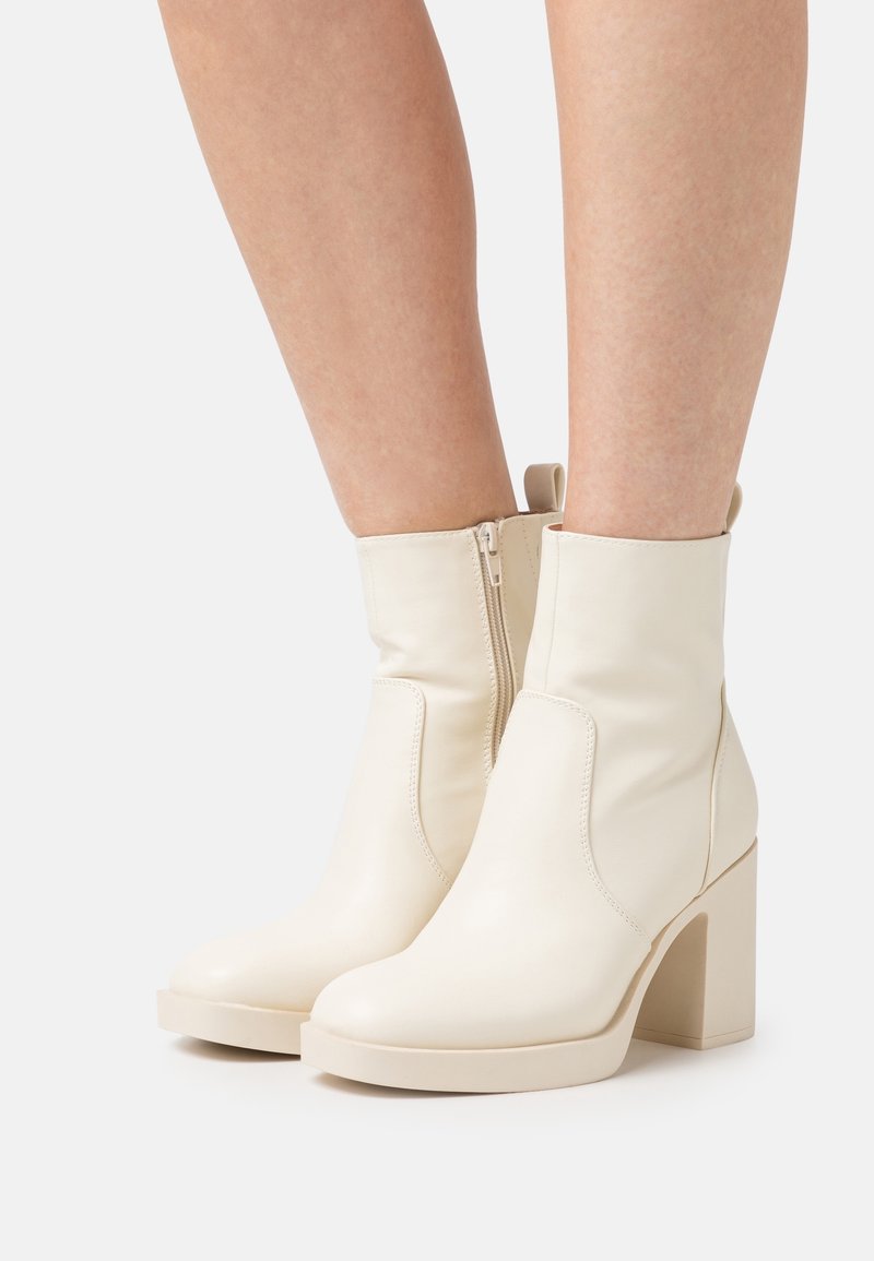 Bottes en cuir couleur crème montant jusqu'au milieu du mollet avec fermetures éclair latérales, talons blocs et languettes d'enfilage, portées sur des jambes nues sur un fond uni.