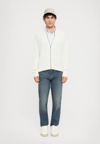 Mladic u bež kapici, s bijelim duksericom na šuplje i svijetloplavoj košulji, plavim farmerkama i bijelim tenisicama, stoji naslonjen na jednostavnu pozadinu.