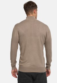 Beige gestrickter Pullover mit langen Ärmeln, hohem Kragen und gerippten Bündchen. Glatte Textur mit figurbetontem Design. Kleines Logo-Detail an der Seite.