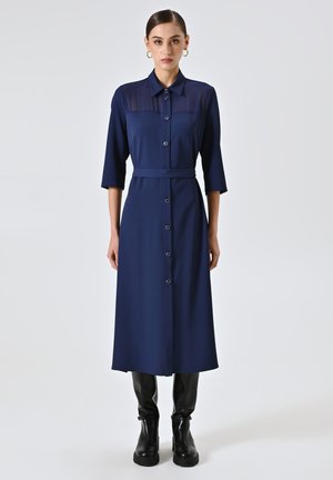 Abito blu navy con bottoni, colletto, maniche a tre quarti e una cintura in vita. Tessuto morbido e silhouette dritta. Stivaletti neri alla caviglia.