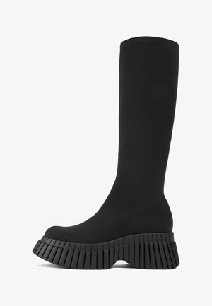 Bota negra hasta la rodilla, hecha de tejido texturizado, con suela gruesa acampanada que presenta estrías verticales y un diseño de punta redondeada.