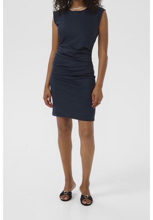 Femme portant une robe bleu marine sans manches jusqu'au genou et des sandales noires à bout ouvert avec des détails de boucle argentée, debout devant un fond uni.