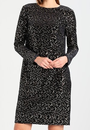 Vrouw met knieproef, lange mouwen zwarte jurk bedekt met zilveren pailletten, staand met handen gevouwen ter hoogte van de taille.