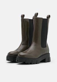 Bottines en cuir olive avec des panneaux élastiques noirs sur les côtés et des semelles en caoutchouc noir épaisses avec une finition texturée. Tirant au sommet.
