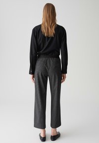Zwarte blouse met lange mouwen en knoopmanchetten, gecombineerd met grijze, wijde broek met een subtiel patroon en elastische tailleband. Zwarte platte schoenen.
