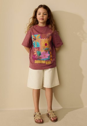 Pige med langt, krøllet hår iført en vinrød T-shirt med print, hvide shorts til knæene og guld sandaler, står op ad en beige væg.