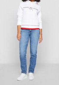 Hvid bomulds-hoodie med "Tommy Hilfiger"-logo, lagdelt over en rød skjorte, kombineret med lyseblå denimjeans og hvide sneakers.