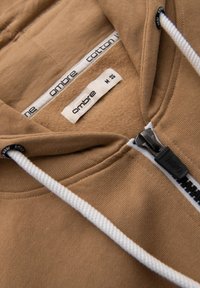 Felpa in cotone marrone con chiusura a zip, cordini bianchi e rivestimento interno in pile. L'etichetta riporta "ombre" con taglia "M."