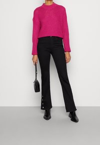 Fuksia neuletakki korkealla kaula-aukolla, yhdistetty mustiin kellotettuihin farkkuihin, joissa on napit nilkan kohdalta. Pitelee pientä mustaa käsilaukkua.