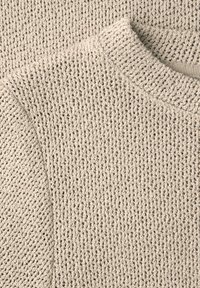 Nærbillede af beige strikket sweater, der viser detaljeret vaffelstrikket tekstur og ribbet rund halsudskæring.