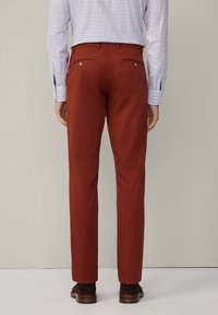 Man in roestkleurige broek, bruin leren schoenen en een wit geruit shirt, staande met zijn gezicht naar weg van een neutrale achtergrond.