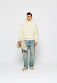 Krēmkrāsas kapučjaka ar kangaroo kabatu, uzvilkta pāri baltai krekls. Gaiši zila džinsa bikses, bēša apavi, tur rokās bēša adīta džemperis.