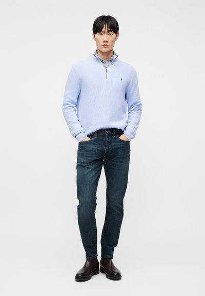 Άνδρας που στέκεται με τα χέρια στις τσέπες, φορώντας ανοιχτό μπλε πουλόβερ με φερμουάρ μέχρι τη μέση, καρό πουκάμισο, σκουρό μπλε τζιν και σκούρα καφέ μπότες.