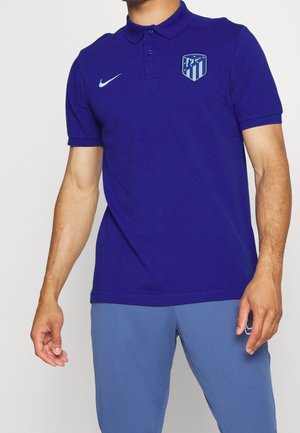Homme portant un polo bleu avec un logo sportif et le Swoosh Nike sur la poitrine, assorti à un pantalon de sport bleu.