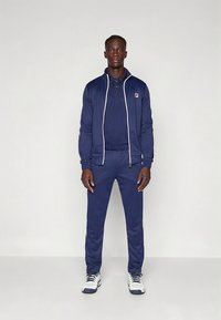 Temno modra trenirka iz gladkega blaga, z jakno z zadrgo in belimi poudarki, polo majico in usklajenimi hlačami. Oblečeno s supergami.