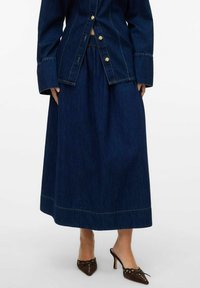 Denim jakna z zlato oblečeni gumbi, v kombinaciji z dolgo temno denim krilo. Obleka ima kontrastne šive in gladko teksturo.