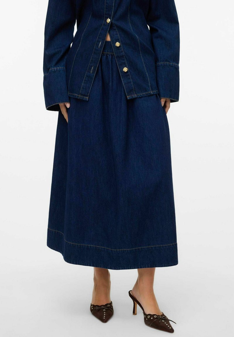 Denim jakna z zlato oblečeni gumbi, v kombinaciji z dolgo temno denim krilo. Obleka ima kontrastne šive in gladko teksturo.