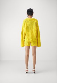 Suéter de punto amarillo con un ajuste holgado, que presenta un patrón texturizado y mangas largas. Combinado con pantalones cortos amarillos a juego.