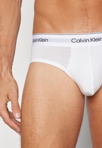 Weiße Baumwollslips mit einem schwarz-weißen Calvin Klein-Logo-Bund, die eine enge Passform und eine glatte Textur zeigen.
