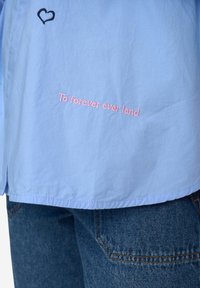 Camicia azzurro chiaro con testo rosa ricamato "To forever ever land" e un cuore nero. Sembra realizzata in un tessuto liscio e leggero.