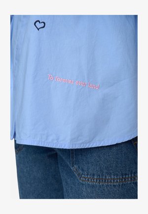 Camicia azzurro chiaro con testo rosa ricamato "To forever ever land" e un cuore nero. Sembra realizzata in un tessuto liscio e leggero.