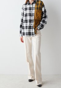 Chemise à carreaux noir et blanc avec une poche patch moutarde, ornée de détails en franges. Associée à un pantalon beige droit et des chaussures noires.
