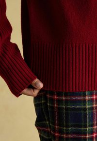 Punane kudumikaelusvöö, millel on ribitud varrukad ja serv, koos ruuduliste pükstega, millel on roheline, sinine, punane ja kollane muster. Ühtlane tekstuur on nähtav.