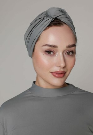 Jeune femme à la peau claire portant un turban gris et un haut à col montant assorti, regardant légèrement sur le côté avec un doux sourire.