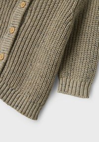 Cardigan en tissu tricoté texturé de couleur beige atténuée avec des éclats marron et bleu. Présente des boutons en bois et des poignets côtelés.