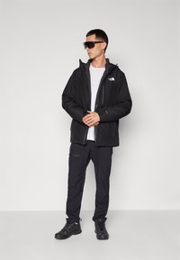Jachetă impermeabilă neagră cu glugă, purtată peste un tricou alb. Asortată cu pantaloni negri și pantofi închiși la culoare. Dispune de un design minimalist și un branding subtil.
