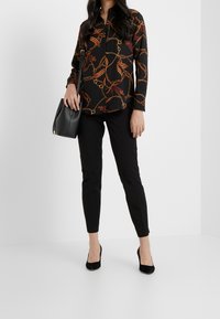 Svart långärmad skjorta med ett kedjemönster i guld och brunt, kombinerad med svarta tajta byxor och svarta högklackade skor; handväska i handen.