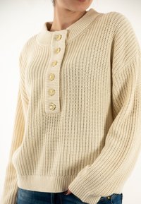 Maglione beige lavorato a maglia con collo ampio e cinque bottoni dorati sulla parte anteriore. Presenta un motivo a coste testurizzato e polsini aderenti.