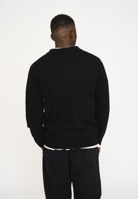 Sort strikket sweater med ribbede kanter og krave, der har en tekstureret overflade. Båret over en hvid skjorte, set fra ryggen mod en ensfarvet baggrund.