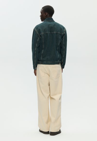 Uomo visto di spalle che indossa una giacca di jeans blu scuro, pantaloni beige a gamba larga e scarpe nere, su uno sfondo chiaro uniforme.