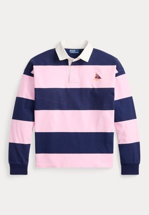 Két ember áll, akik ugyanolyan rózsaszín és tengerészkék csíkos rögbi pólókat viselnek; az egyik bézs nadrággal és fehér cipővel, a másik bézs rövidnadrággal, tengerészkék tornacipővel, fehér sapkával és napszemüveggel párosítja.