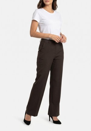 Femme portant un t-shirt blanc uni à manches courtes, un pantalon droit marron foncé et des chaussures à talons hauts noires, debout les mains jointes.
