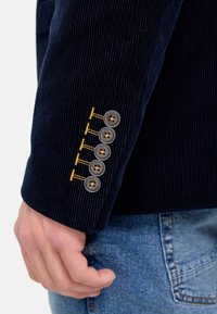 Navy cordfløyelsblazer med erme som har fire grå knapper og gulltråd detaljer, som viser teksturert stoff og skreddersydd passform.